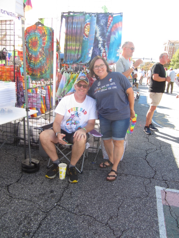 At an outdoor pride festival are two smiling PFLAG parents — a seated man in sunglasses and a Free Dad Hugs shirt, being embraced by a woman wearing a shirt that says "Be careful who you hate, it could be someone you love." Colorful tie-dyed Pride t-shirts and banners are displayed on a rack behind them.