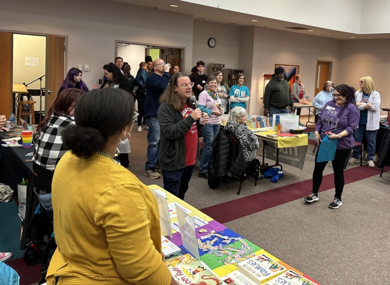 A person in a red shirt makes an announcement during the Queer Book Festival. PFLAG's table is seen in the background.