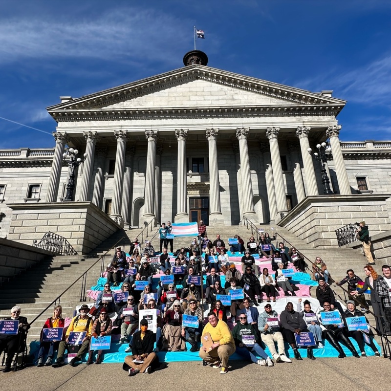 A large group of people is seated on the steps of the South Carolina Statehouse, which have been draped in fabric in the colors of the transgender flag. Many are holding up signs that say "Trans youth belong" and "Don't disrupt trans lives."